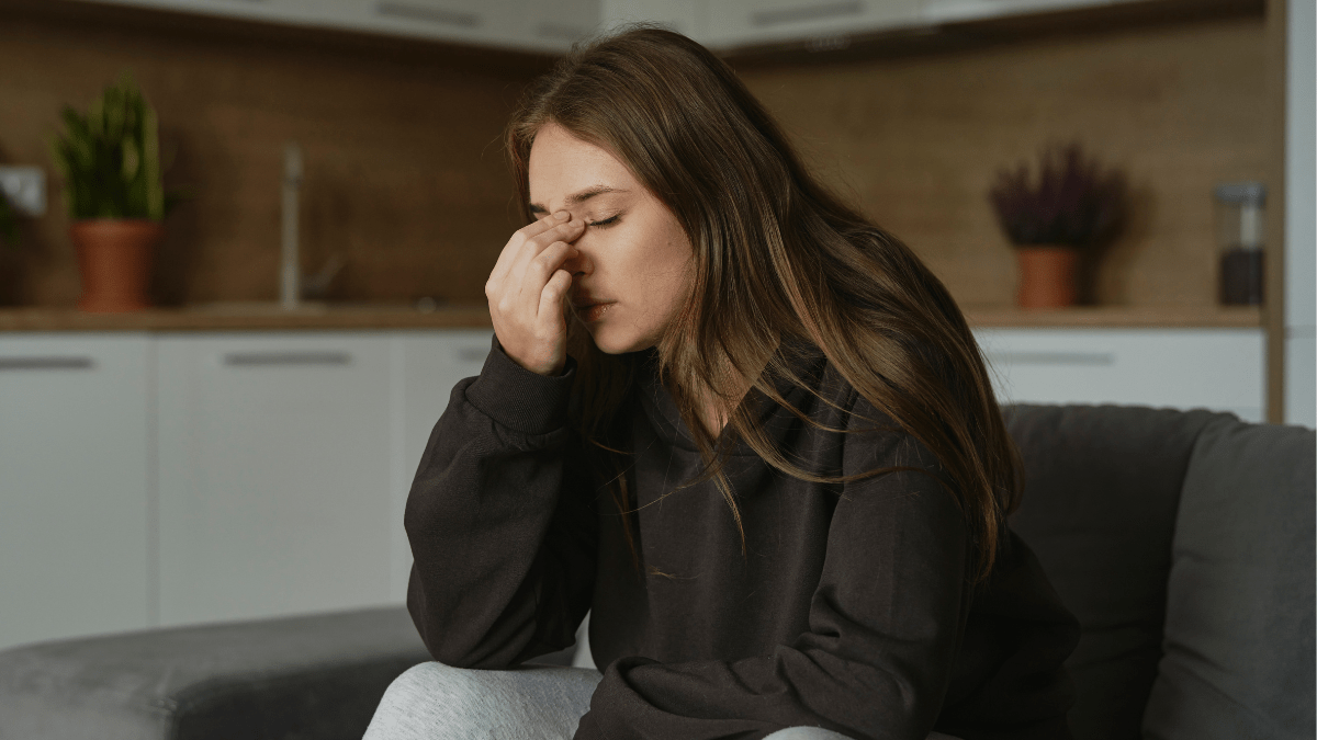 Eine junge Frau mit braunen Haaren sitzt auf einer dunklen Couch und hält eine Hand an ihre Nase.