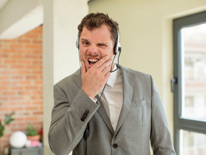 Ein Mitarbeiter in einem Callcenter mit Headset verzieht wÃ¤hrend eines Telefonats genervt das Gesicht.