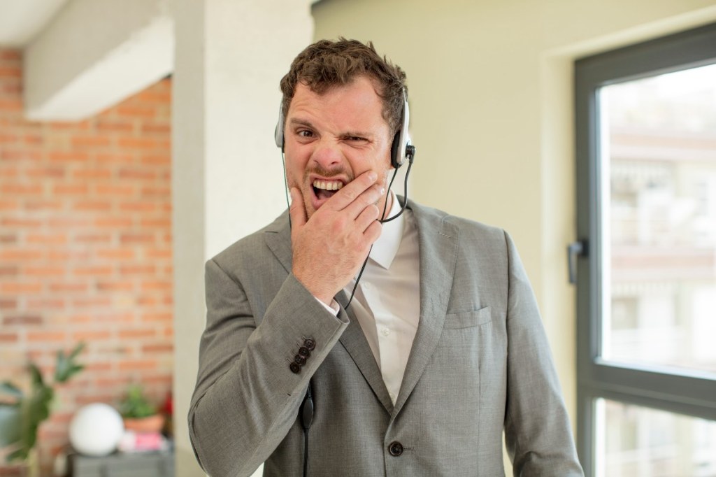 Ein Mitarbeiter in einem Callcenter mit Headset verzieht während eines Telefonats genervt das Gesicht.