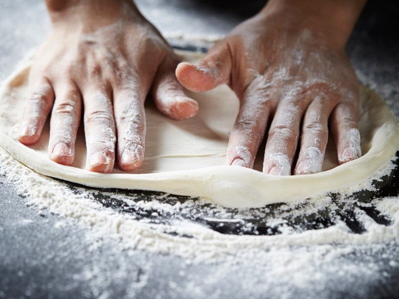 Ein Pizzateig wird auf einer mit Mehl bestäubten Unterlage geknetet.