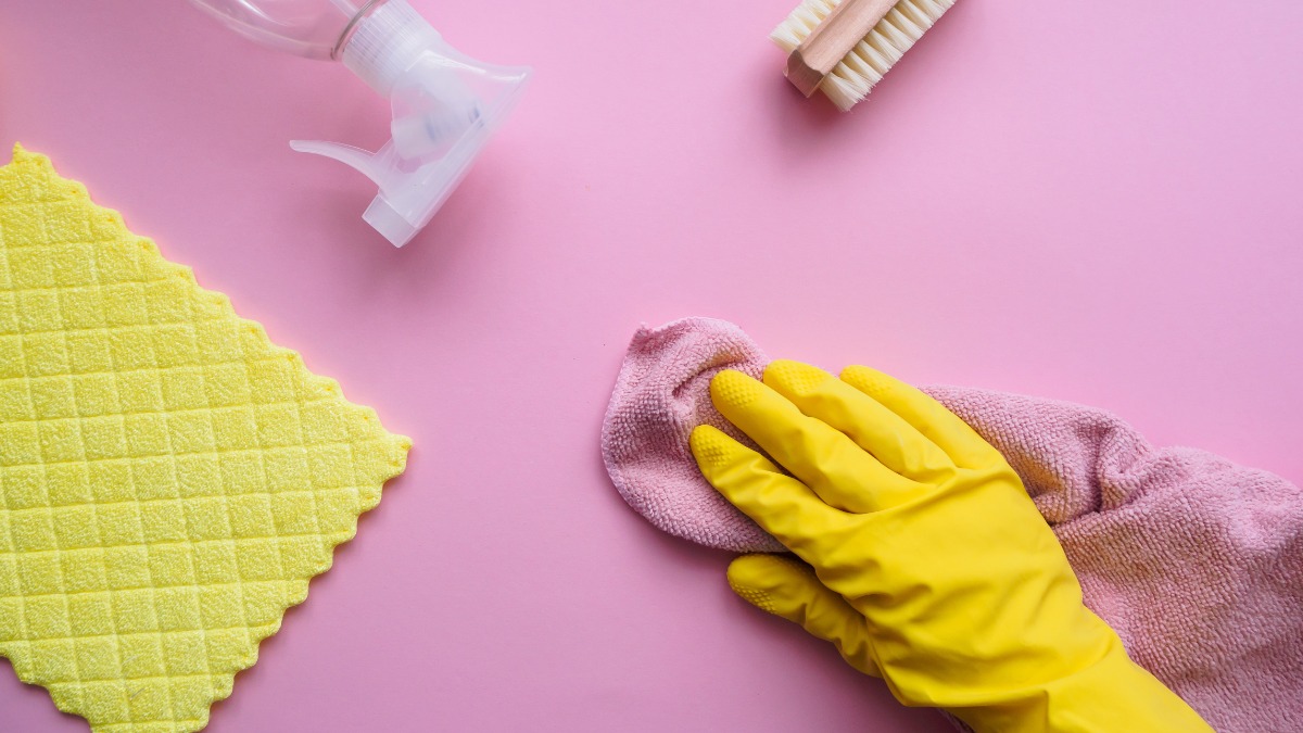 Utensilien zum Putzen und eine Hand mit einem gelben Gummihandschuh auf einem rosa Hintergrund