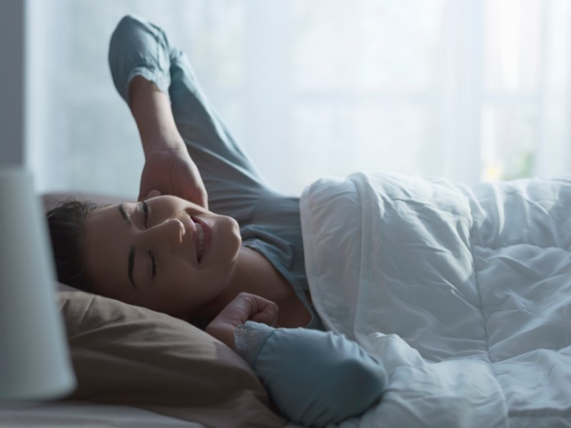 Eine junge Frau, die gerade aufgewacht ist, liegt im Bett und streckt sich gutgelaunt.