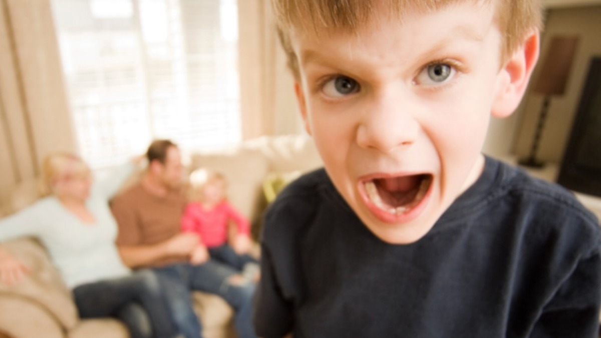 Kleiner Junge schreit in die Kamera, während hinter ihm seine Familie auf dem Sofa sitzt