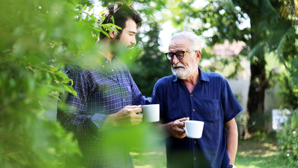 Ein älterer und ein jüngerer Mann unterhalten sich im Freien. Sie halten beide eine weiße Kaffeetasse in der Hand.