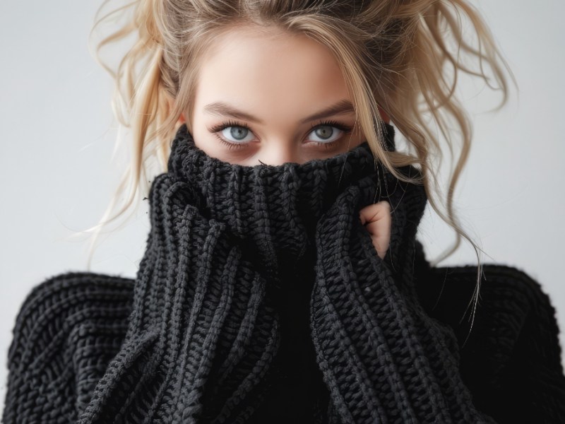 Eine junge, blonde Frau versteckt sich vor Scham im Rollkragen ihres Pullovers.