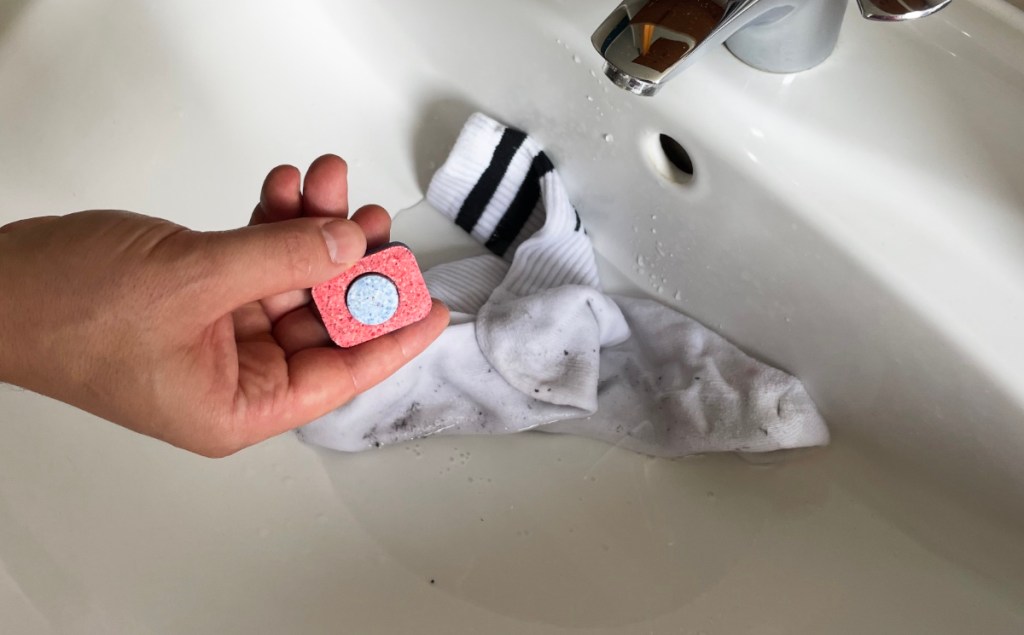 Eine Hand hält einen Spülmaschinen-Tab über eine weiße Socke, die in einem Waschbecken einweicht.
