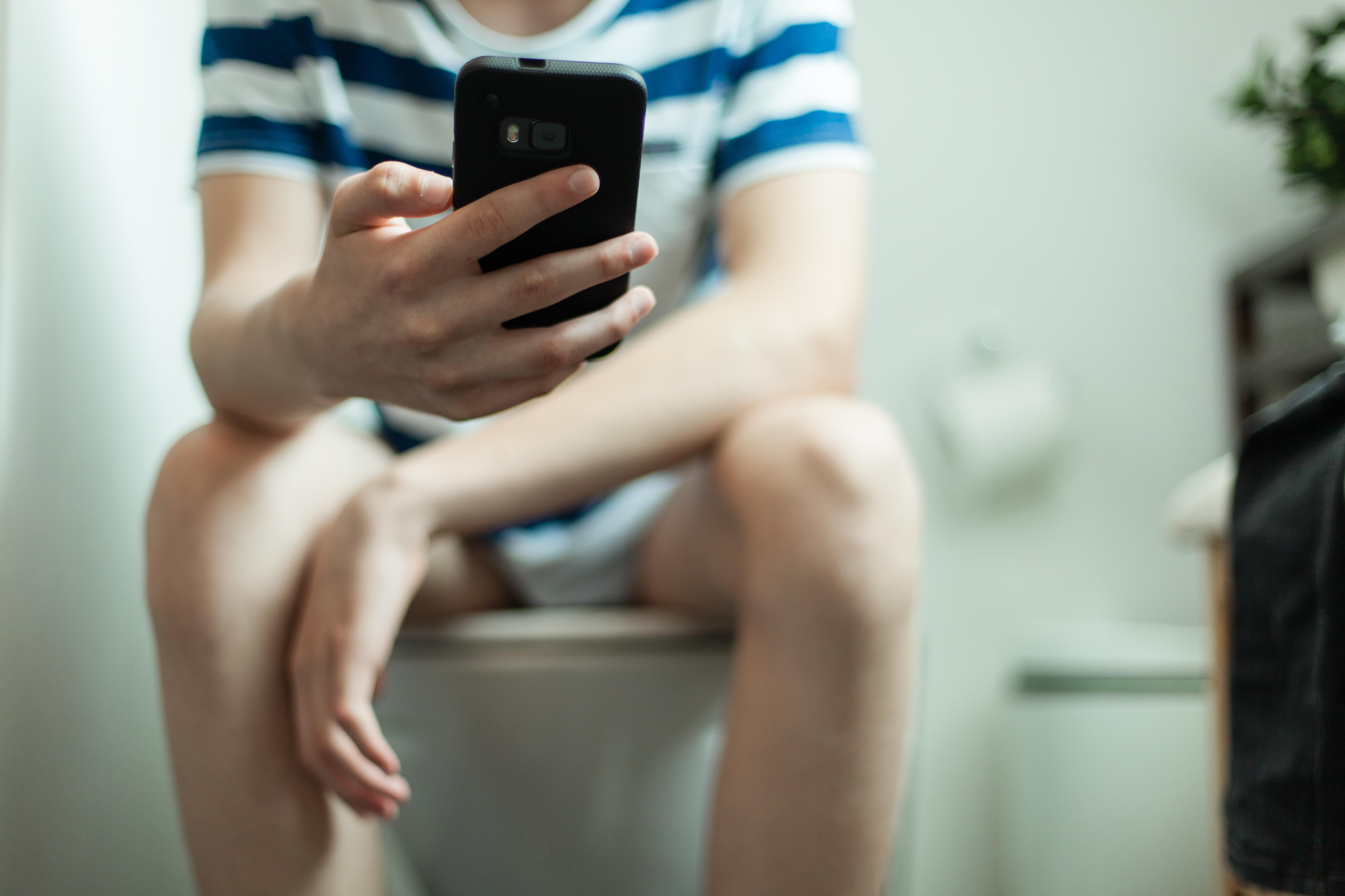 Ein Mann sitzt auf einer Toilette und hat ein Smartphone in der Hand.