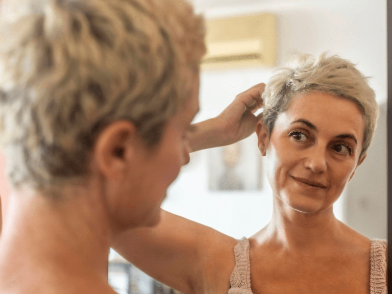 Eine Frau mit kurzen grauen Haaren steht vor einem Spiegel.