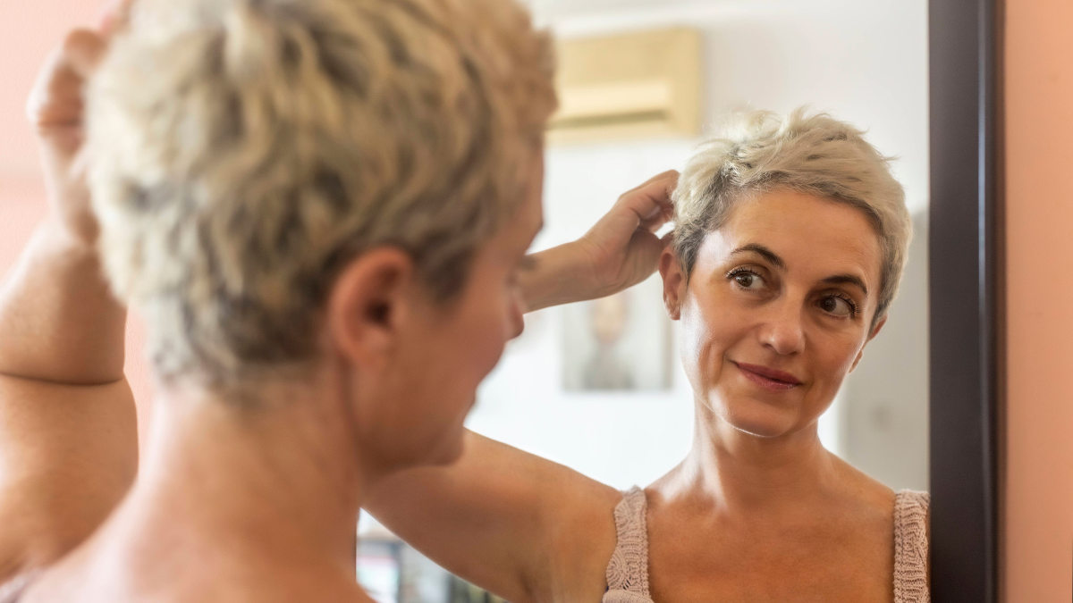 Eine Frau mit kurzen grauen Haaren steht vor einem Spiegel.