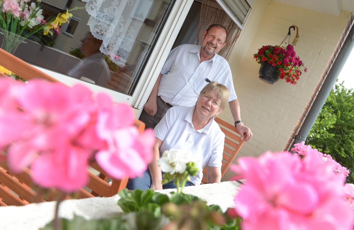 Ein Rentner-Ehepaar posiert hinter pinken Geranien auf dem Balkon für ein Foto.