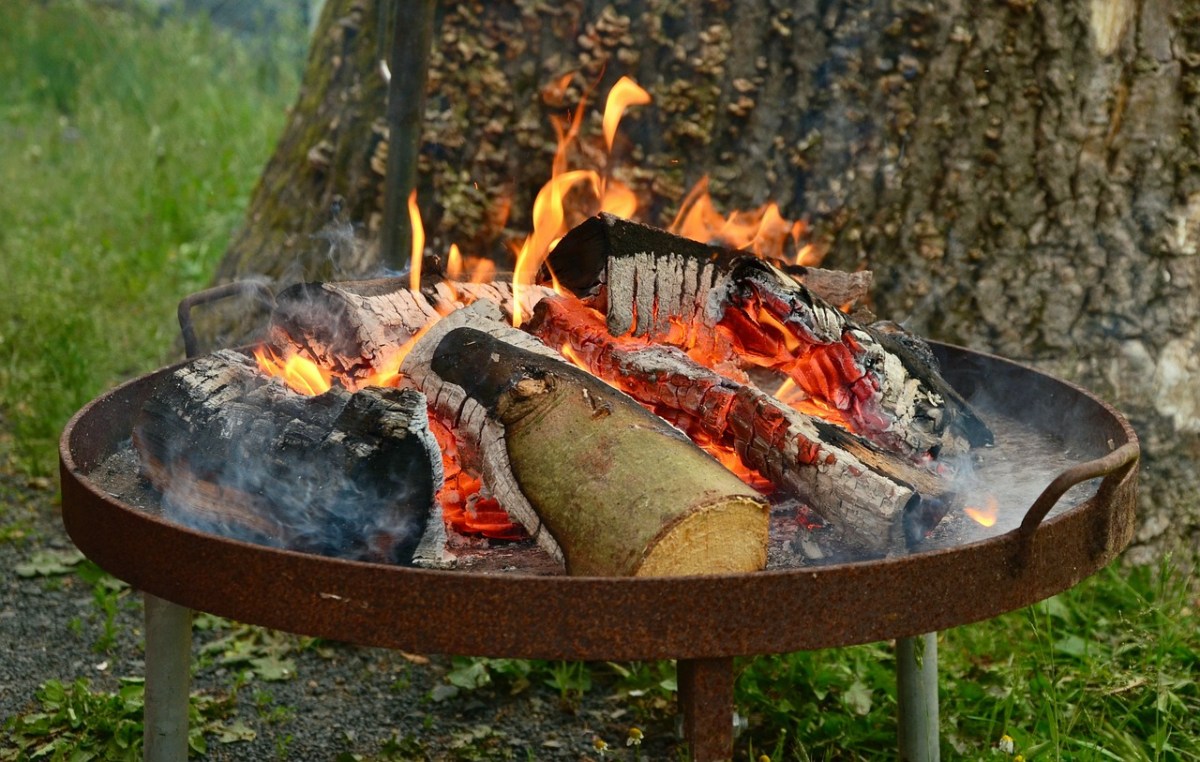 Eine Feuerschale, in der ein Feuer brennt, steht in einem Garten vor einem Baum.