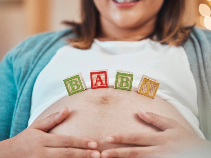 Schwangere Frau hält ihre Hände am Bauch, auf diesem stehen Würfel mit dem Wort Baby.