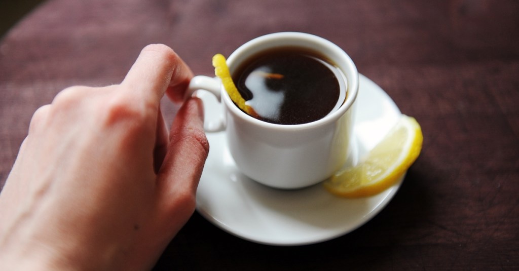 Jemand hält eine Tasse Espresso mit Zitrone in der Hand und setzt zum Trinken an.