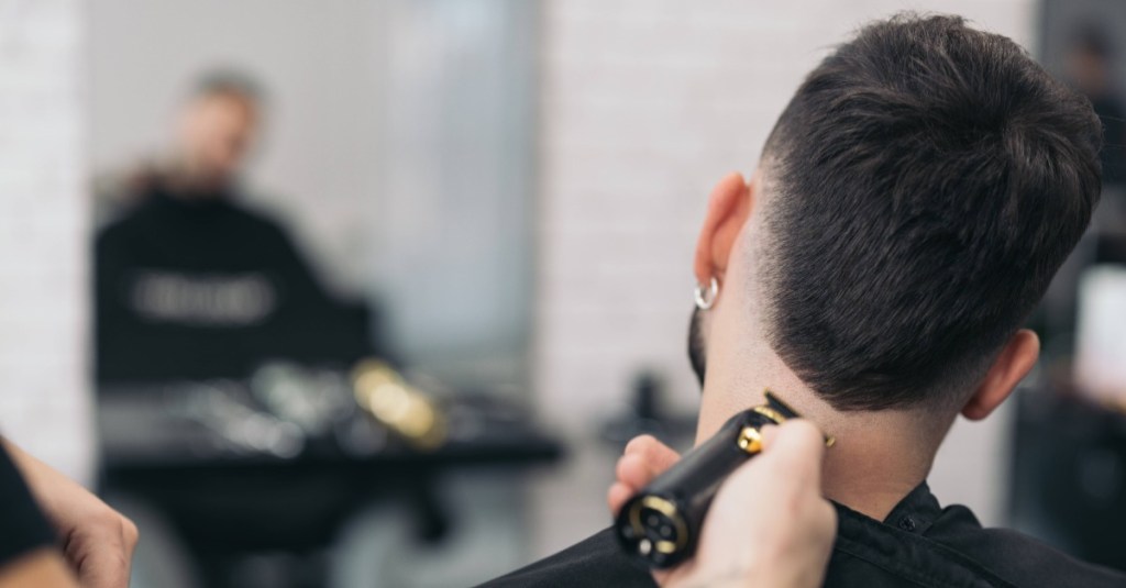 Ein junger Mann sitzt auf einem Friseurstuhl während der Frisuer mit einer Haarschneidemaschine die Haare kürzt.