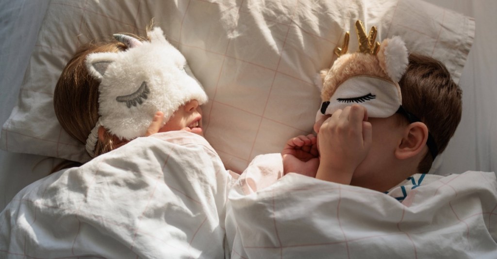 zwei Kinder liegen mit Schlafmasken im Bett