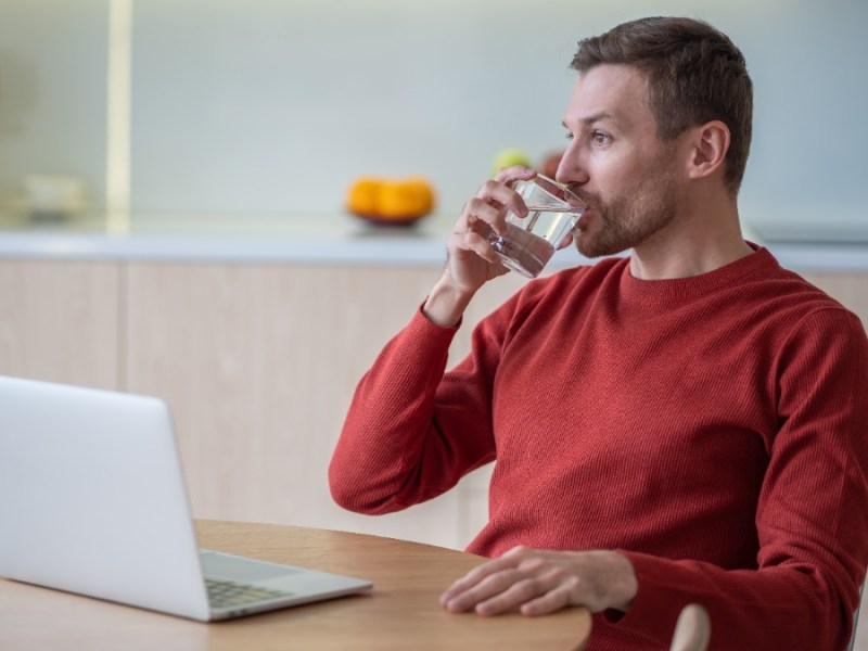 Ein Mann sitzt vor seinem Laptop und trinkt ein Glas Wasser