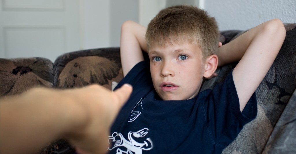 Junge sitzt mit verschränkten Armen auf der Couch, während eine Hand auf ihn zeigt