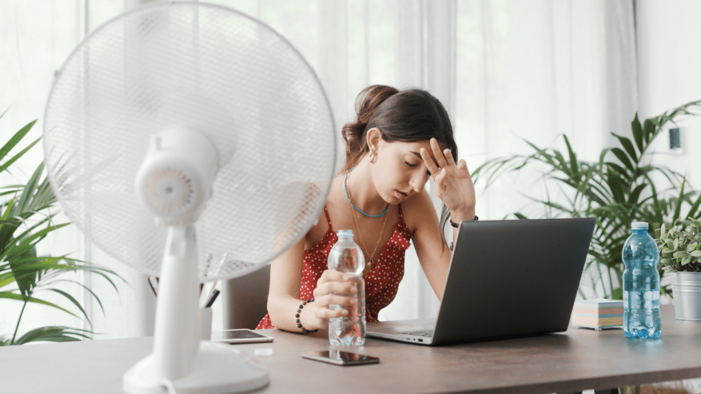 Eine Frau stützt sich auf ihren Schreibtisch und hält eine Wasserflasche in der Hand. Im Vordergrund sieht man einen weißen Ventilator.