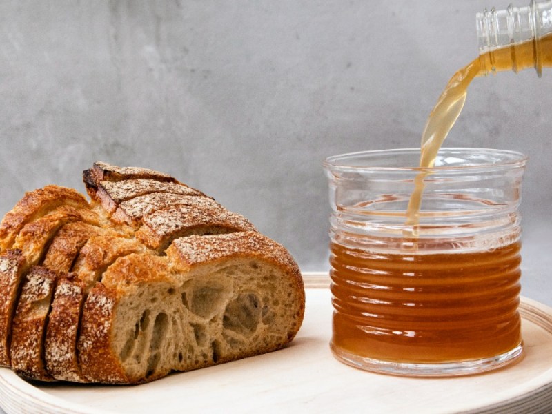 Ein Laib Brot und daneben steht ein Glas, in das eine bräunliche Flüssigkeit gegossen wird.