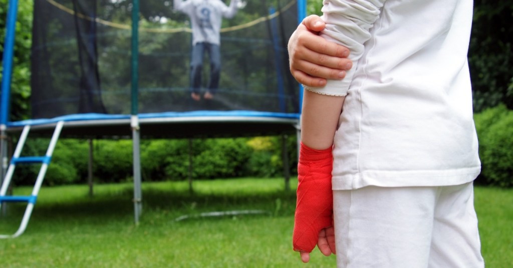 Ein Mädchen mit einem gebrochenen Finger steht vor einem Trampolin, auf dem ein Junge hüpft