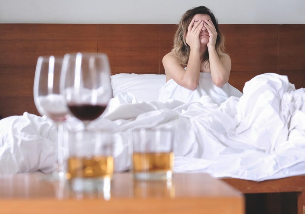 Junge Frau im Bett, daneben Gläser mit Wein und Alkohol.