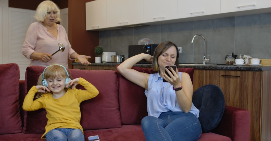 Mutter und Tochter sitzen auf dem Sofa, während die Oma im Hintergrund schimpft