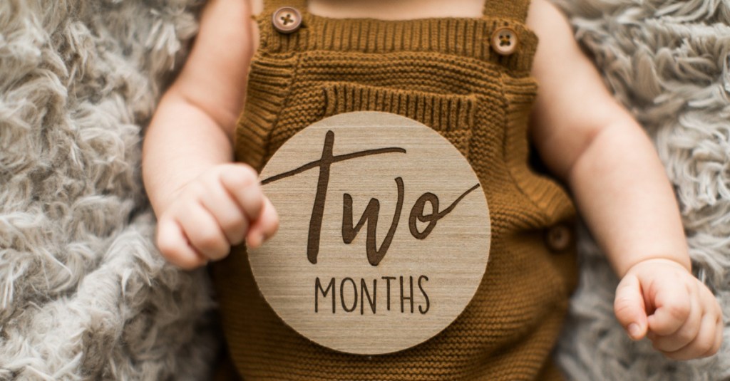 kleiner Junge in einem Overall, auf seinem Bauch liegt ein Holzkreis mit der Aufschrift "two month"