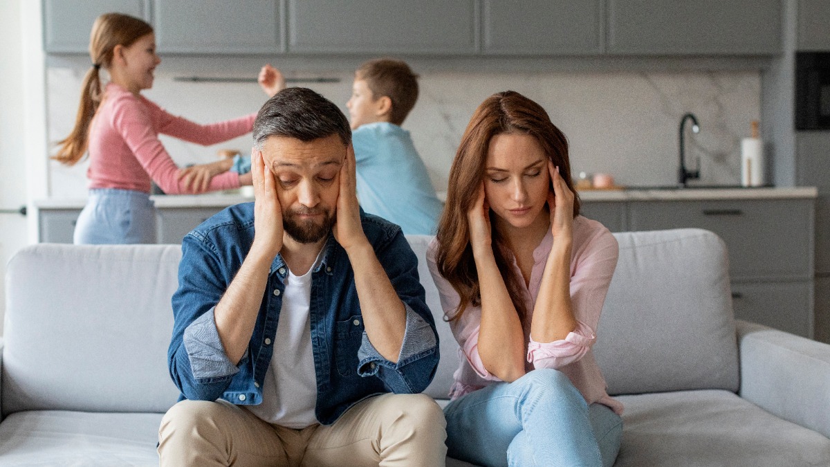 Eltern sitzen genervt auf der Couch, während sich die Kinder im Hintergrund streiten