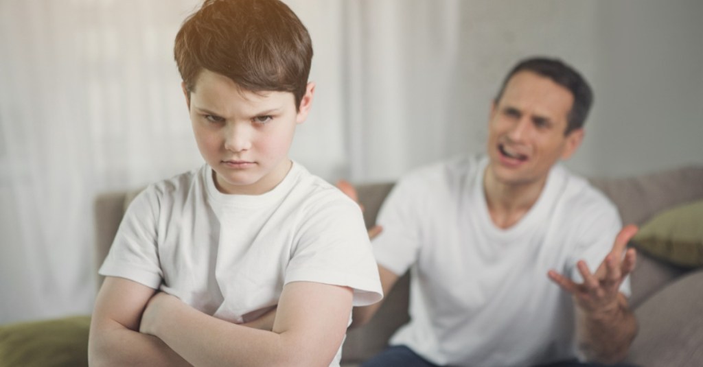 Junge guckt mit verschränkten Armen in die Kamera, während sein Vater im Hintergrund verärgert auf ihn einredet