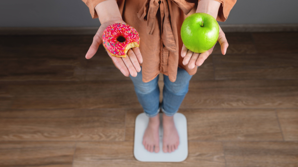 Eine Frau steht barfuß auf der Waage und hält in einer Hand einen grünen Apfel und in der anderen ein Donut.