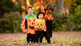 Drei Kinder mit Kürbiskostümen auf einer Wiese mit buntem Herbstlaub