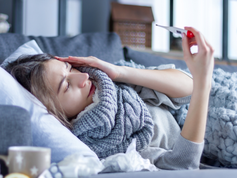 Eine Frau liegt krank auf dem Sofa und schaut auf ein Fieberthermometer.