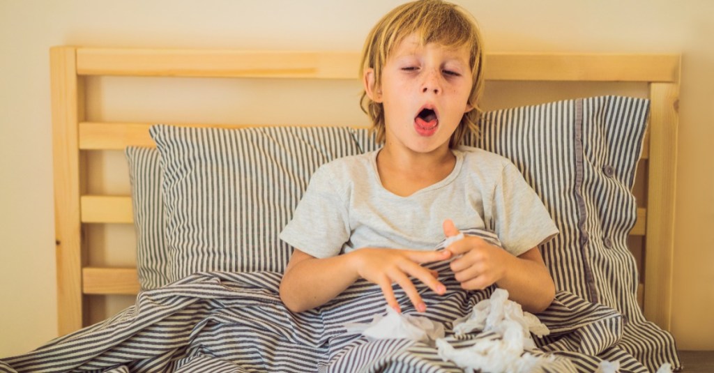 kranker Junge sitzt im Bett, um ihn verteilt liegen Taschentücher