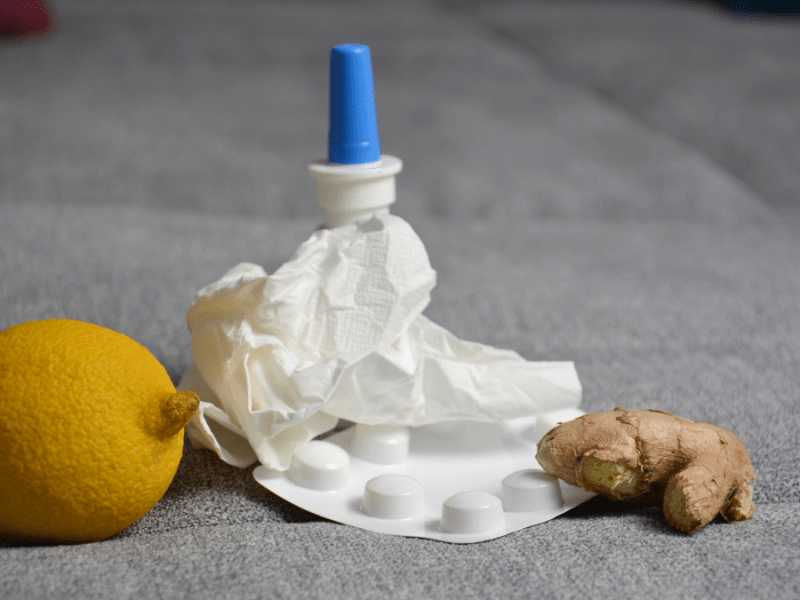 Eine Zitrone, ein Nasenspray, ein Medikamentenblister, ein zerknÃ¼lltes Taschentuch und ein StÃ¼ck Ingwer liegen auf einer grauen Couch.