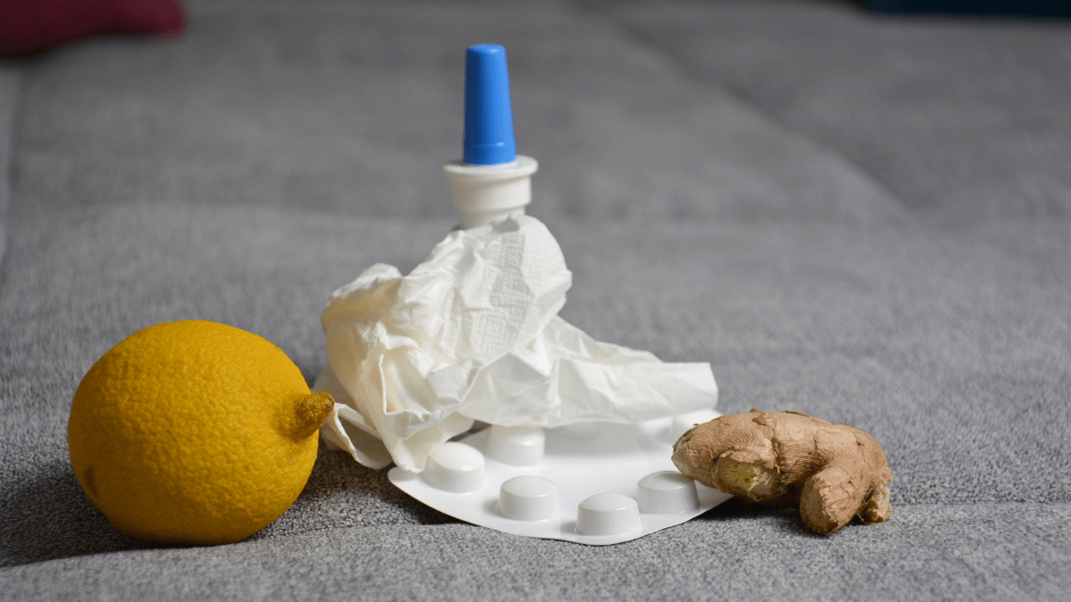 Eine Zitrone, ein Nasenspray, ein Medikamentenblister, ein zerknülltes Taschentuch und ein Stück Ingwer liegen auf einer grauen Couch.