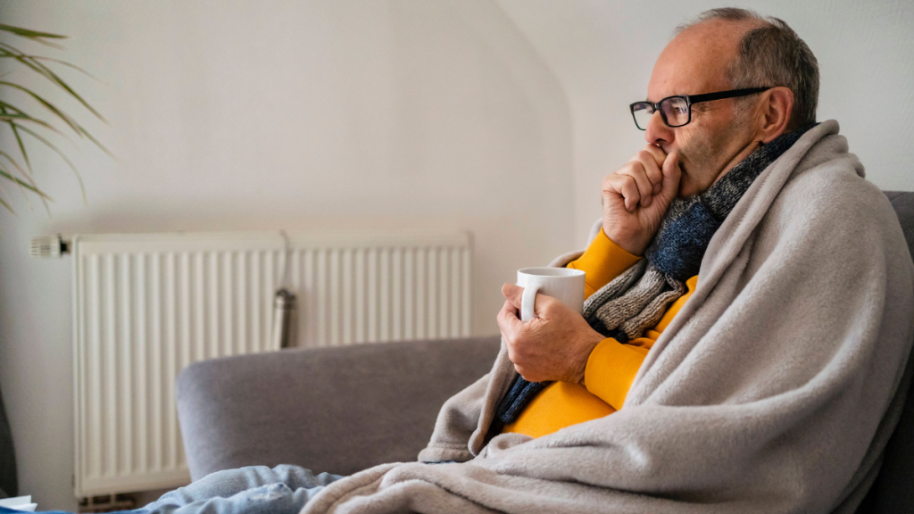 Ein Mann sitzt in eine Decke gehüllt, mit einer Tasse Tee auf dem Sofa.