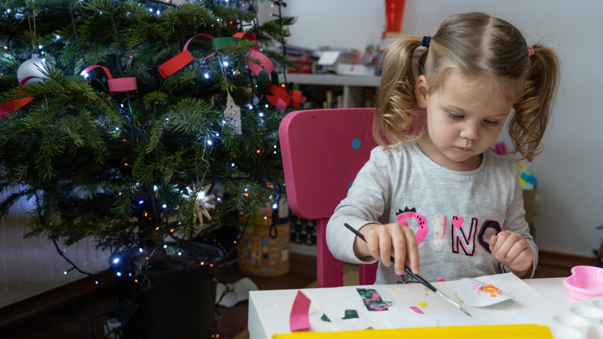 nÃ¼tzliche Geschenke Kinder: ein kleines MÃ¤dchen sitzt neben einem Weihnachtsbaum am Tisch und bastelt