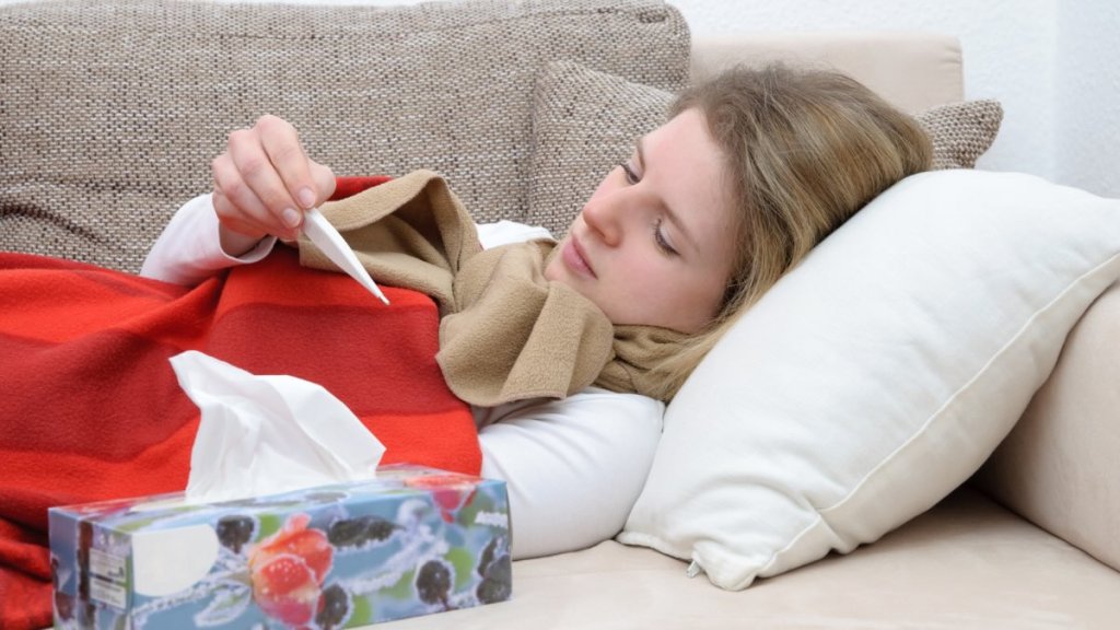 Eine Frau liegt auf der Couch und schaut auf ein weißes Fieberthermometer.