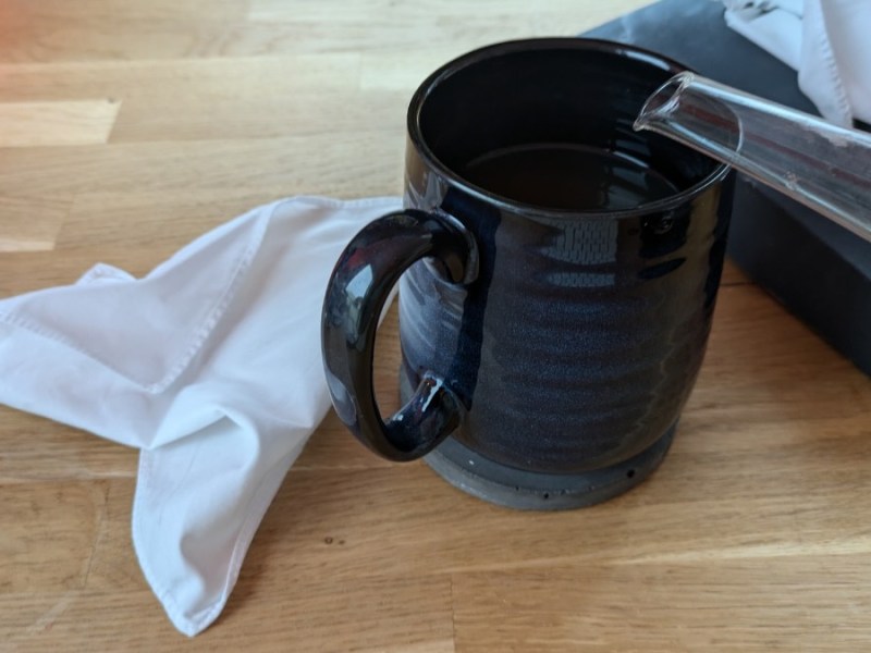 Eine blaue Teetasse, in die Tee eingegossen wird. Daneben liegt ein zerknülltes Taschentuch.