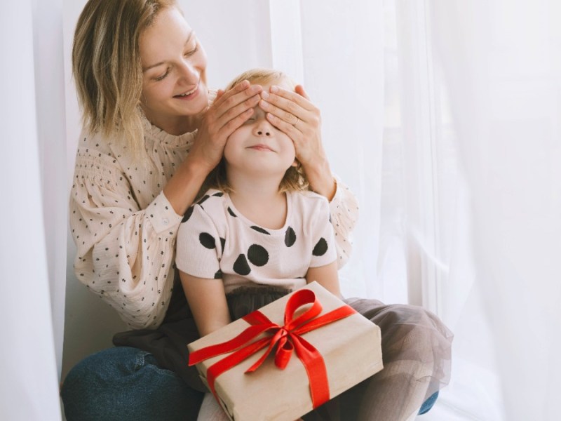 Alleinerziehend an Weihnachten: Mutter verdeckt die Augen ihrer Tochter, auf deren Schoß ein Geschenk liegt