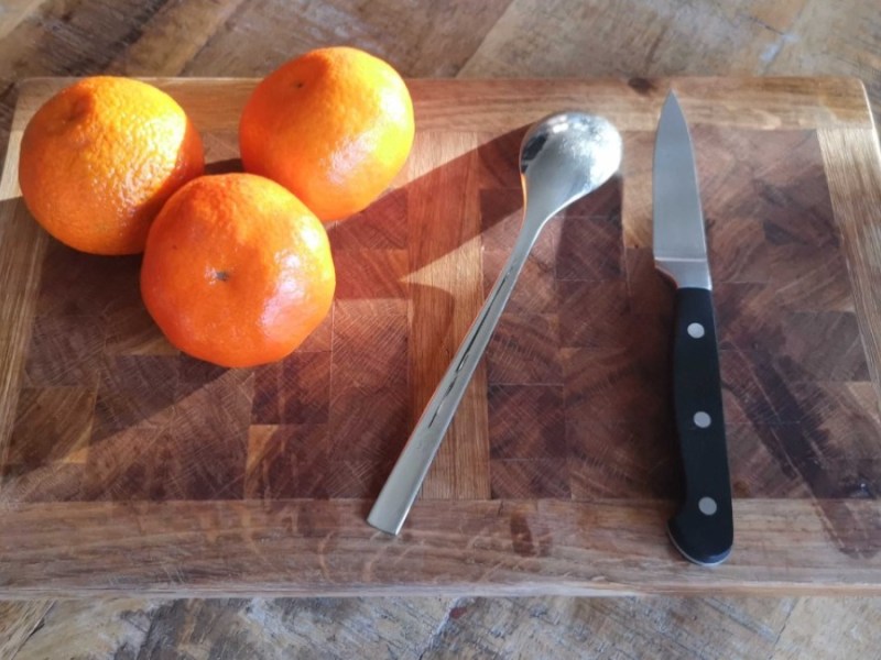 Mandarinen schälen: drei Mandarinen, ein Esslöffel und ein Messer liegen auf einem Holzbrett