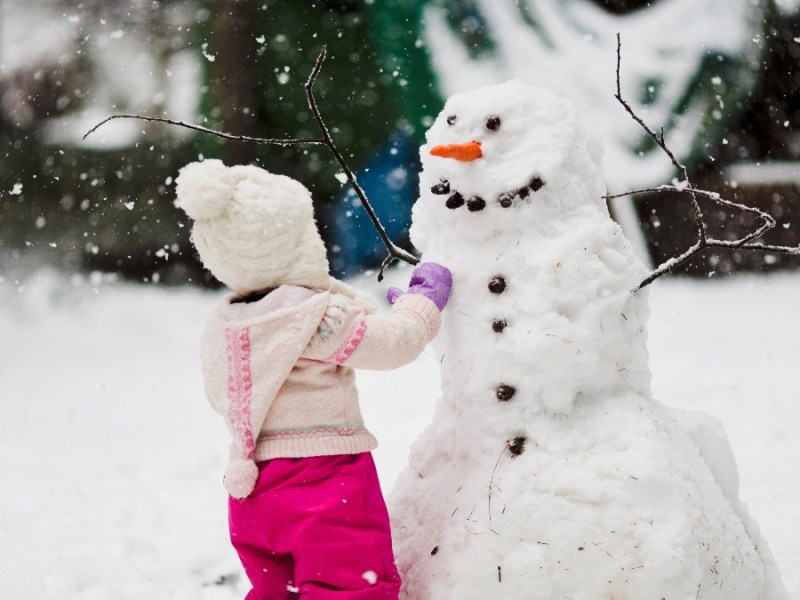 Schneemann bauen: ein kleines Mädchen steht vor einem Schneemann