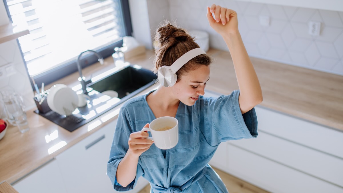 Eine Frau hat KopfhÃ¶rer auf und eine Tasse in der Hand und tanzt in ihrer KÃ¼che.