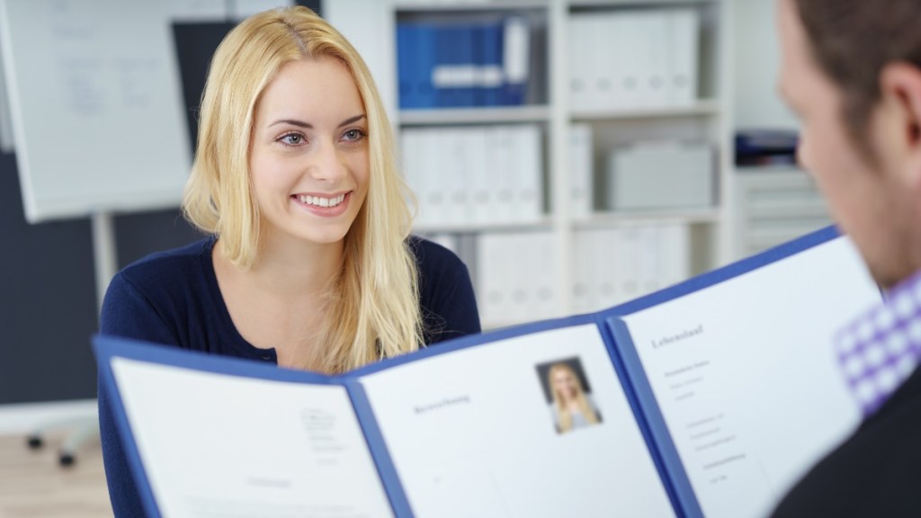 Eine blonde junge Frau sitzt vor einer Person, die ihre Bewerbungsmappe anschaut.