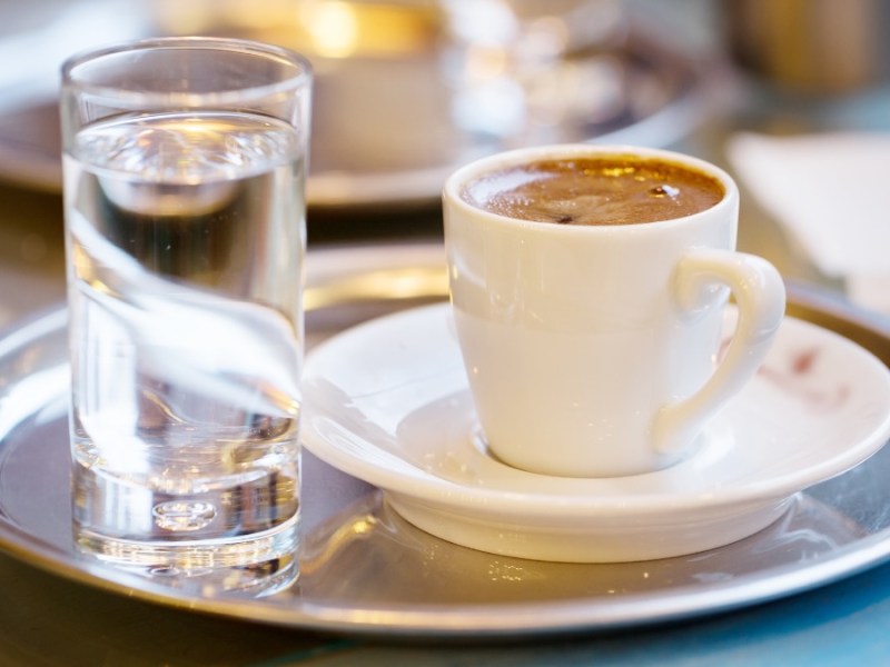 Ein silbernes Tablett mit einem Espresso und einem Glas Wasser stehen auf einem Tisch.